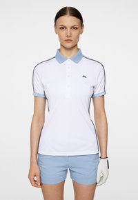 Vit polo skjorta med ljusblå krage och ärmdetaljer, prydd med svarta detaljer och en logotyp på vänster bröst. Kombineras med ljusblå shorts.