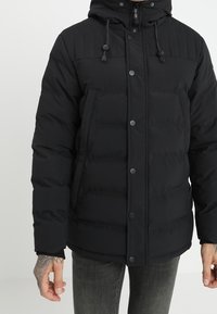 Svart kviltad pufferjacka med huva och tryckknappar, bärs av en person med tatuerad hand och grå jeans.