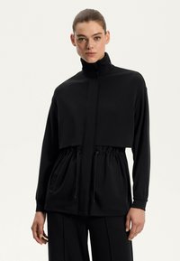Zwarte ritsjack met hoge kraag, verkort ontwerp, elastische taille en voorzakken. De stof lijkt soepel en lichtgewicht.