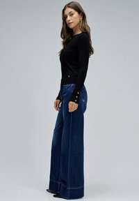 Haut noir à manches longues associé à un jean en denim foncé à taille haute avec des jambes larges. Le haut présente un logo discret, tandis que le jean a une finition soignée et des bas de jambes prononcés.
