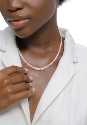 Collier de perles composé de petites perles blanches rondes et brillantes, posé sur une peau foncée et lisse, accompagné d'une chemise de couleur claire.