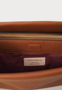 Brown textured leather handbag interior with gold zipper, featuring a maroon lining and a label stating "Coccinelle, Authentic Italian Leather."