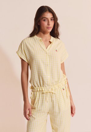 Femme portant un haut à manches courtes boutonné à carreaux vichy jaune et blanc et un pantalon assorti avec de petites cerises brodées sur la poitrine et la taille.