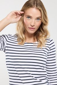 Blond kvinde med blå øjne iført en hvid og sort stribet langærmet skjorte, der rører ved sit hår med den ene hånd mod en ensfarvet baggrund.