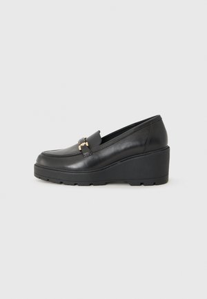 Mocassins pretos de salto em cunha feitos de couro liso com ponta arredondada, apresentando um detalhe decorativo em metal dourado na parte da frente.