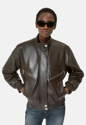 Femme portant des lunettes de soleil noires en forme d'œil de chat et une veste en cuir marron foncé avec poches zippées, mains dans les poches, sur fond blanc.