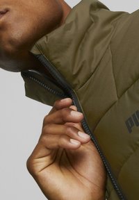 Doudoune vert olive avec col montant et fermeture éclair. Présente une texture matelassée et des détails de fermeture éclair noirs. Logo de la marque visible sur le tissu.