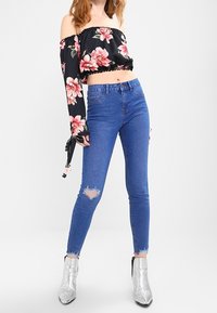 Svart blommig off-shoulder kort topp med volangkant, kombinerad med blå skinny jeans med slitna knän och ankellång fåll.