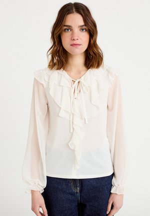 Hvid bluse med flæsekant ved halsudskæringen og ærmekanter, med bindebåndslukning, lavet af transparent stof med en blød tekstur, båret over mørke jeans.