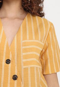 Camicetta gialla senape con strisce verticali bianche, maniche corte, scollatura a V profonda e taschino frontale. Presenta bottoni neri per la chiusura.