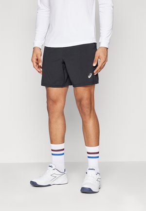 Mand iført sorte sportsshorts, hvid langærmet skjorte, hvide sokker med blå og røde striber og hvide sportssko.