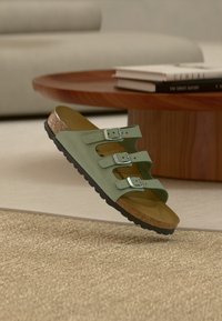 Πράσινο σανδάλι με τρεις λουρίδες και ρυθμιζόμενες ασημένιες αγκράφες, φαρδύ πάτο από φελλό και μαύρη υφασμάτινη σόλα από καουτσούκ, κρεμασμένο πάνω από μία υφαντή επιφάνεια.