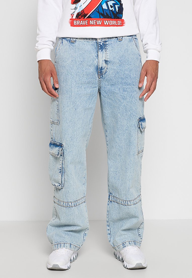 Jeans cargo azzurri, dal fit largo, con tasche laterali e una texture di denim sbiadito. Indossati con una felpa bianca con grafica e sneakers bianche.
