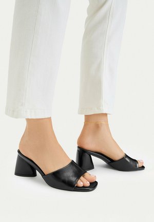 Pies con zapatos tipo mule negros abiertos, con tacón cuadrado, combinados con pantalones blancos recortados y una delgada pulsera de tobillo dorada en un tobillo.