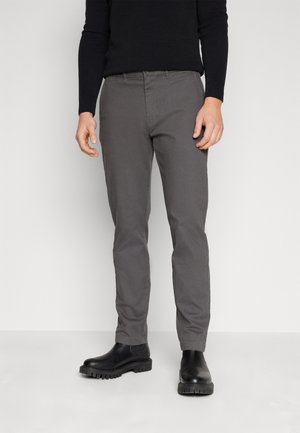 Uomo con pantaloni grigi a gamba dritta, camicia nera a maniche lunghe e stivaletti neri con suola spessa, in piedi su uno sfondo neutro.