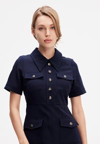 Robe bleu marine avec un col, des manches courtes, deux poches avant et des détails en boutons argentés sur le devant. Texture de tissu lisse.