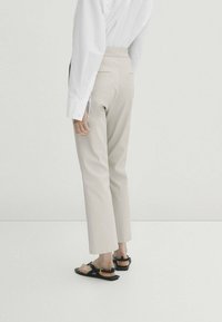 Pantalon beige à coupe droite, avec deux poches arrière et une taille haute, associé à des sandales plates noires. Chemise blanche à manches longues portée.