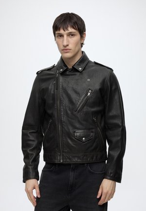 Homme aux cheveux bruns courts portant un blouson de motard en cuir noir avec détails de zip et de poches, debout sur un fond blanc uni.