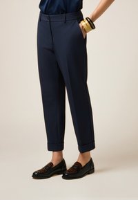 Donna che indossa pantaloni su misura navy a lunghezza caviglia, scarpe mocassino a righe nere e marroni, con una mano in tasca e bracciali impilati sul polso.