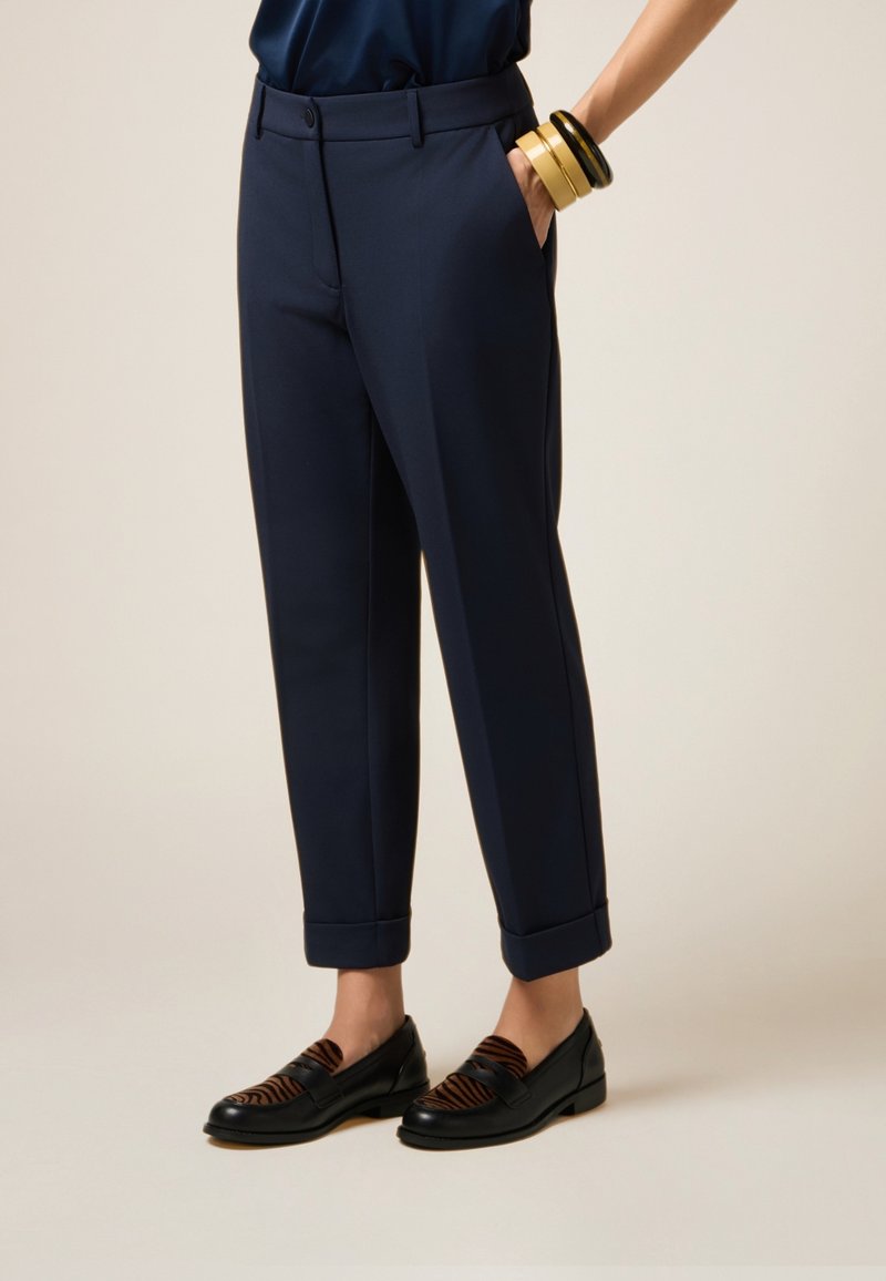Donna che indossa pantaloni su misura navy a lunghezza caviglia, scarpe mocassino a righe nere e marroni, con una mano in tasca e bracciali impilati sul polso.