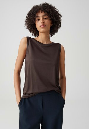 Femme aux cheveux bouclés portant un haut sans manches marron foncé et un pantalon bleu marine, debout devant un fond clair uni, les mains dans les poches.
