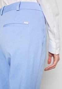 Pantalon bleu clair sur mesure avec une poche arrière et un logo BOSS en métal blanc, associé à un poignet de chemise blanc à manches longues et une main.