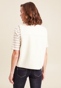 Cremfarbener Fleece-Westen mit abgerundetem Saum, getragen über einem gestreiften T-Shirt. Verfügt über eine Rückenpasse und ein ärmelloses Design. Dunkle Jeans vervollständigen das Outfit.