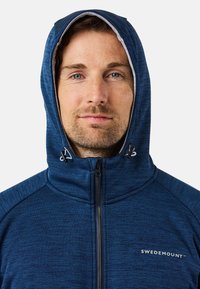 Felpa con zip di colore blu scuro, con colletto alto, cappuccio regolabile e dettagli di colore blu più chiaro. Presenta un logo sul petto e un tessuto strutturato.