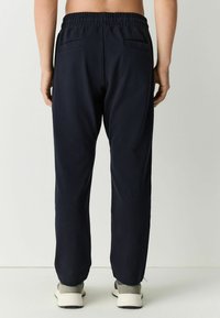 Pantalones de chándal azul marino con una cinturilla elástica, dos bolsillos traseros, piernas ajustadas y una textura suave. Se presentan con zapatillas de color claro.