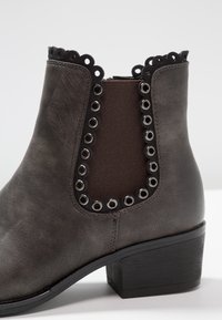 Szare botki za kostkę o strukturalnym wykończeniu, z elastycznym bocznym panelem, ozdobnymi przelotkami i niskim, blokowym obcasem. Detaile z falbany na górnej krawędzi.