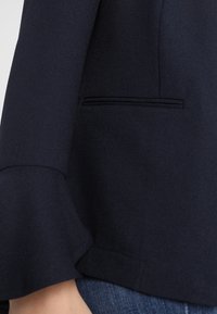 Námořnická modrá bunda s tesnějším střihem, jemnou texturou a standardní kapsou s klopou na boku.