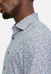 Chemise à boutons avec un fond blanc orné de motifs tourbillonnants complexes en bleu, rouge et noir. Col court, texture du tissu lisse.