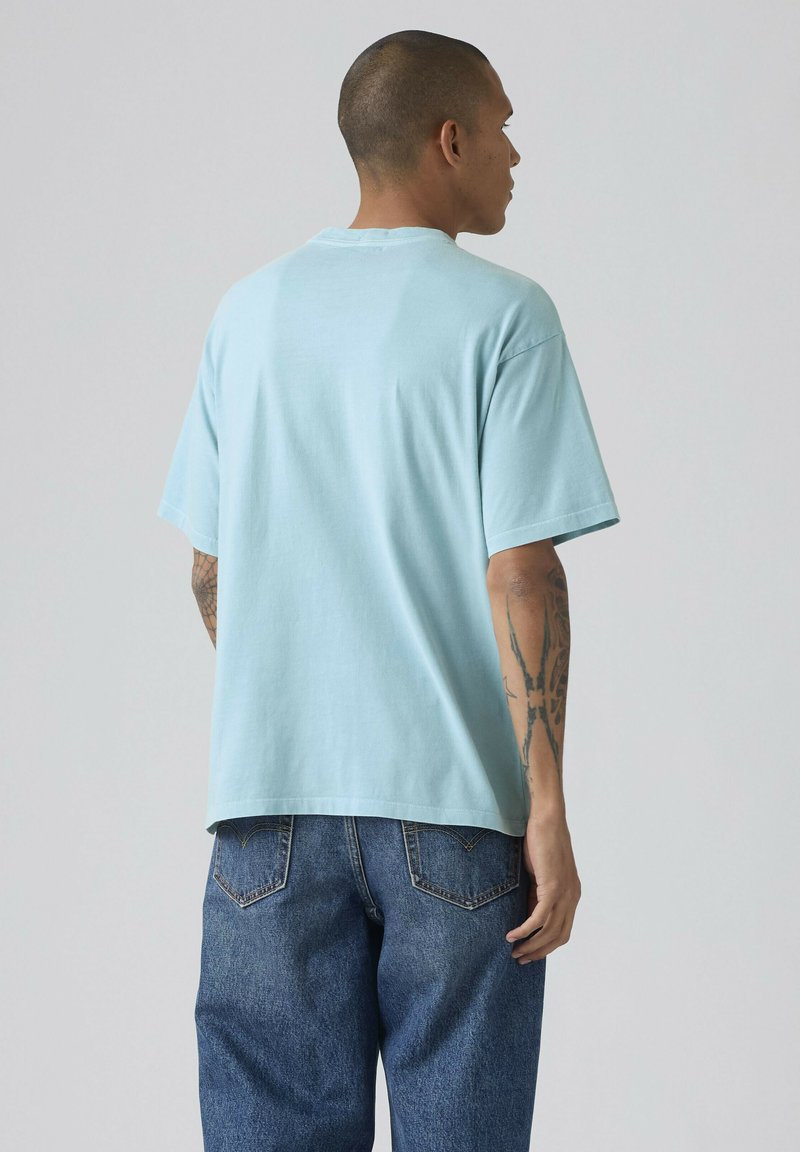 Hombre con brazos tatuados que lleva una camiseta azul claro y vaqueros azules, de pie mirando hacia atrás contra un fondo gris liso.