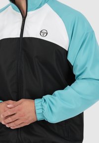 Zwarte zip-up jas met lichtblauwe mouwen en witte details, voorzien van elastische manchetten en een klein logo op de borst.