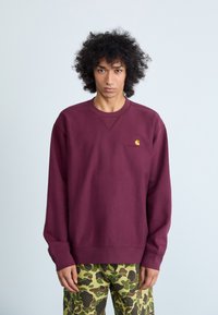 Bordový svetr s kulatým výstřihem, oversize střih a žebrované manžety. Na levé straně hrudi má malé žluté logo. Texturovaná tkanina.