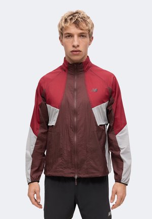Junger Mann mit hellen lockigen Haaren trägt eine Jacke mit Reißverschluss in Rot, Braun und Weiß sowie schwarze Sportshorts, steht vor einem schlichten Hintergrund.