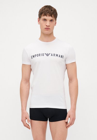 T-shirt en coton blanc avec un graphique "Emporio Armani" bleu marine. Conception ajustée avec des manches courtes, porté avec un short en boxer noir.