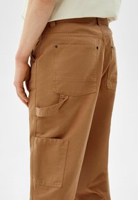 Brązowe spodnie bojówki o luźnym kroju, z wieloma kieszeniami, widocznymi szwami i gładką teksturą bawełny.