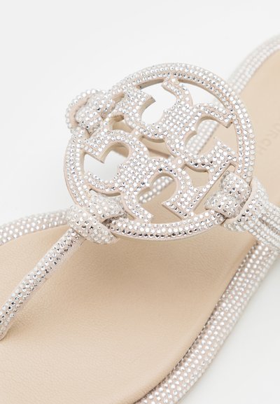Sandale thong embellizate cu argint, având o piesă decorativă circulară cu design decupat, bretele texturate și o talpă netedă bej.