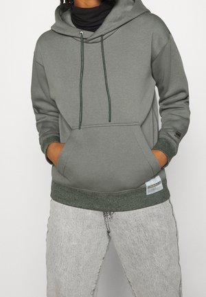 Personne portant un sweat à capuche gris-vert avec poche avant et cordons, associé à un jean denim gris clair, les mains dans la poche.
