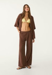 Camicia oversized marrone a coste con bottoni abbinata a pantaloni a gamba larga dello stesso colore e un bikini giallo. Sandali neri piatti completano il look.
