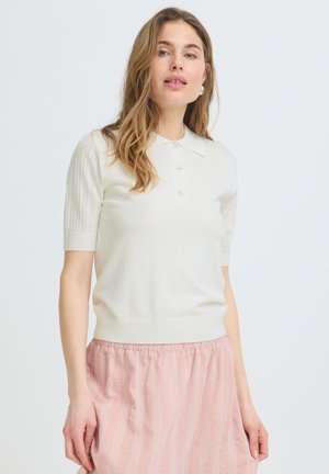 Žena s dugom valovitom kosom, odjevena u bijelu pletenu majicu kratkih rukava s gumbima i ružičastu prugastu suknju na jednobojnoj pozadini.