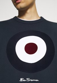 Personne portant un sweat-shirt foncé avec un grand motif circulaire en forme de cible rouge, blanc et noir sur la poitrine et le texte « Ben Sherman » en dessous.