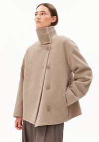 Beige wool coat with a high collar, featuring five buttons on the front and wide, structured sleeves. Smooth texture and relaxed fit.