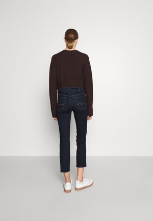 Kvinde med brunt hår bundet tilbage iført mørk sweater, mørke korte jeans og hvide sneakers, står vendt mod en ensfarvet hvid væg.