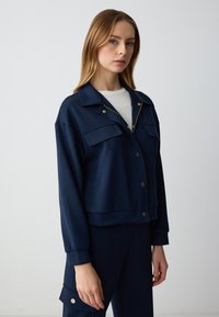 Giacca a zip blu navy realizzata in tessuto liscio, con due tasche frontali, hardware color oro e un design cropped.