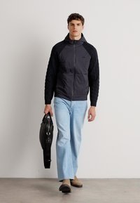 Casaco preto com fecho éclair e mangas texturizadas, jeans azul claro de perna reta, sapatos castanhos e uma mala de couro preta. Design simples e moderno.