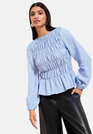 Hellblaue Bluse mit langen Ärmeln, geraffter Textur und Schößchen-Saum, mit elastischen Bündchen und rundem Ausschnitt. Schwarze Lederhose.