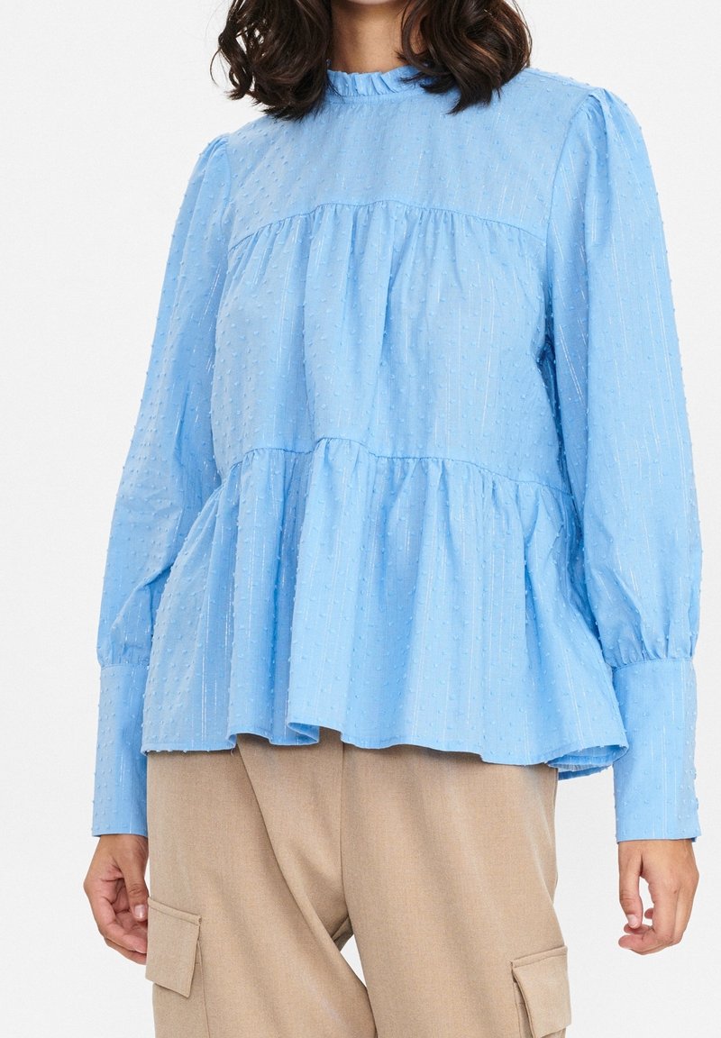 Blusa azzurra chiara con tessuto testurizzato, orlo arricciato e maniche lunghe. Presenta un collo alto e sottili strisce verticali nel materiale.
