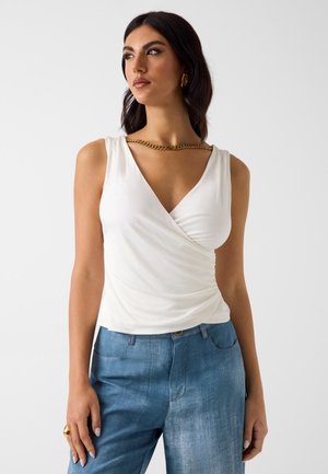 Femme portant un haut cache-cœur blanc sans manches, un jean bleu, un collier chaîne en or, des boucles d'oreilles créoles en or et une bague en or, debout devant un fond blanc.